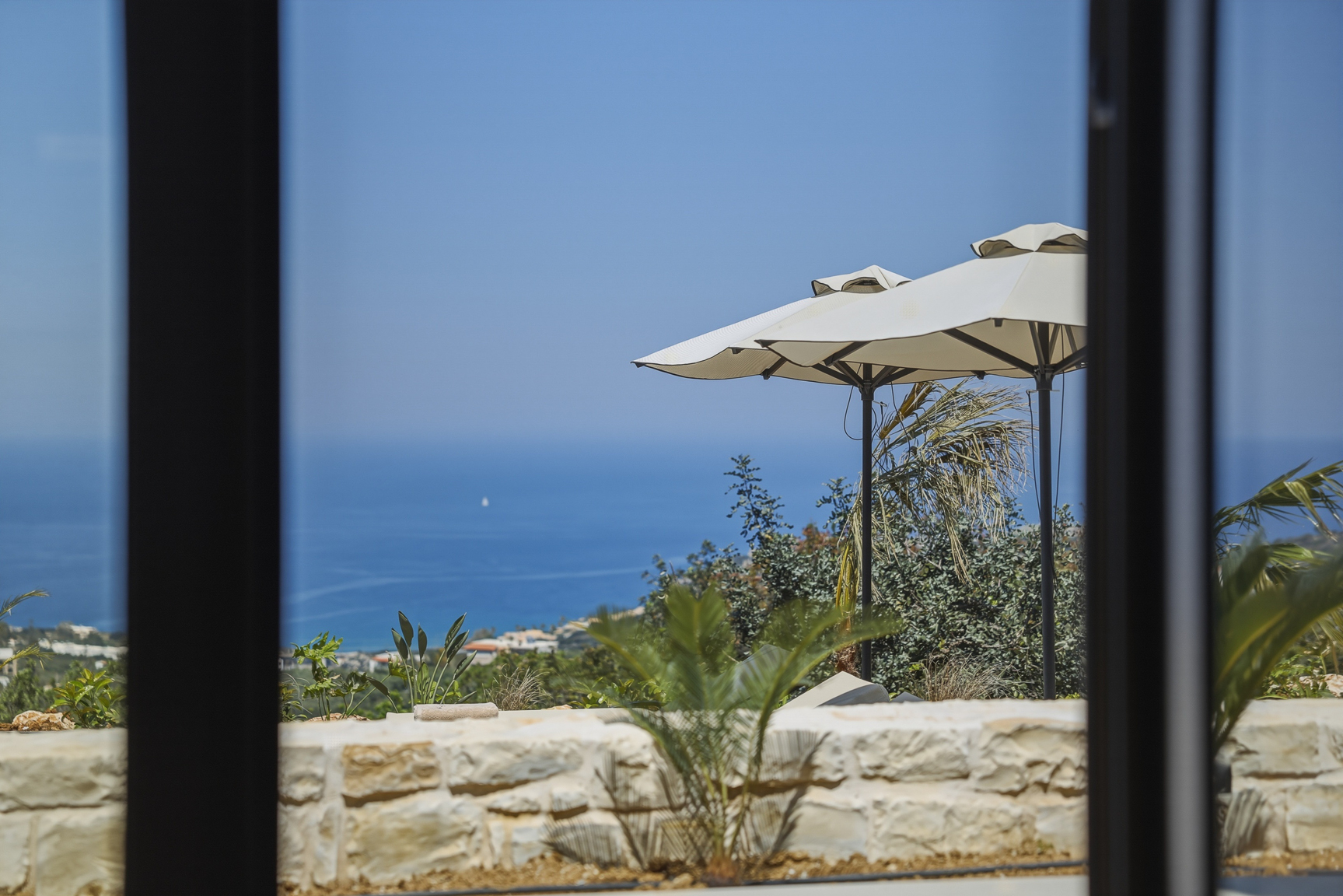 Blick durch das Panoramafenster der Villa Selene über Sonnenschirme und Steinmauer zum libyschen Meer